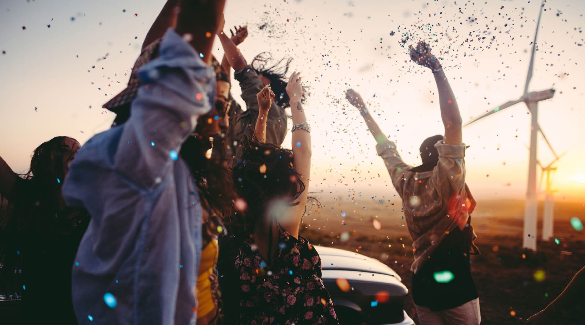 Group of multi-ethnic teens dressed in boho style partying outdoors throwing colourful confetti in a wind farm at sunset during their american carefree road trip